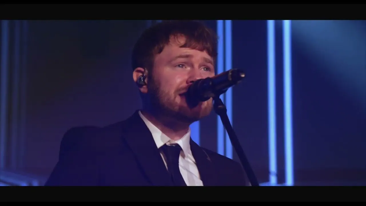 Man with short brown hair and beard wearing a suit and tie singing into a microphone on stage with blue vertical lights in the background.