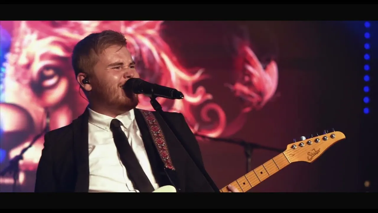 Musician in a black suit and white shirt singing into a microphone while playing an electric guitar on stage with a colorful lion face backdrop.