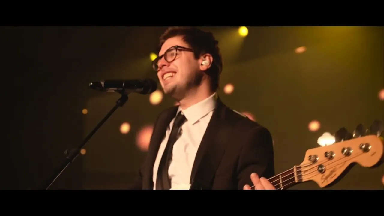 Smiling musician in a suit and glasses singing into a microphone while playing a bass guitar on stage with warm lighting.