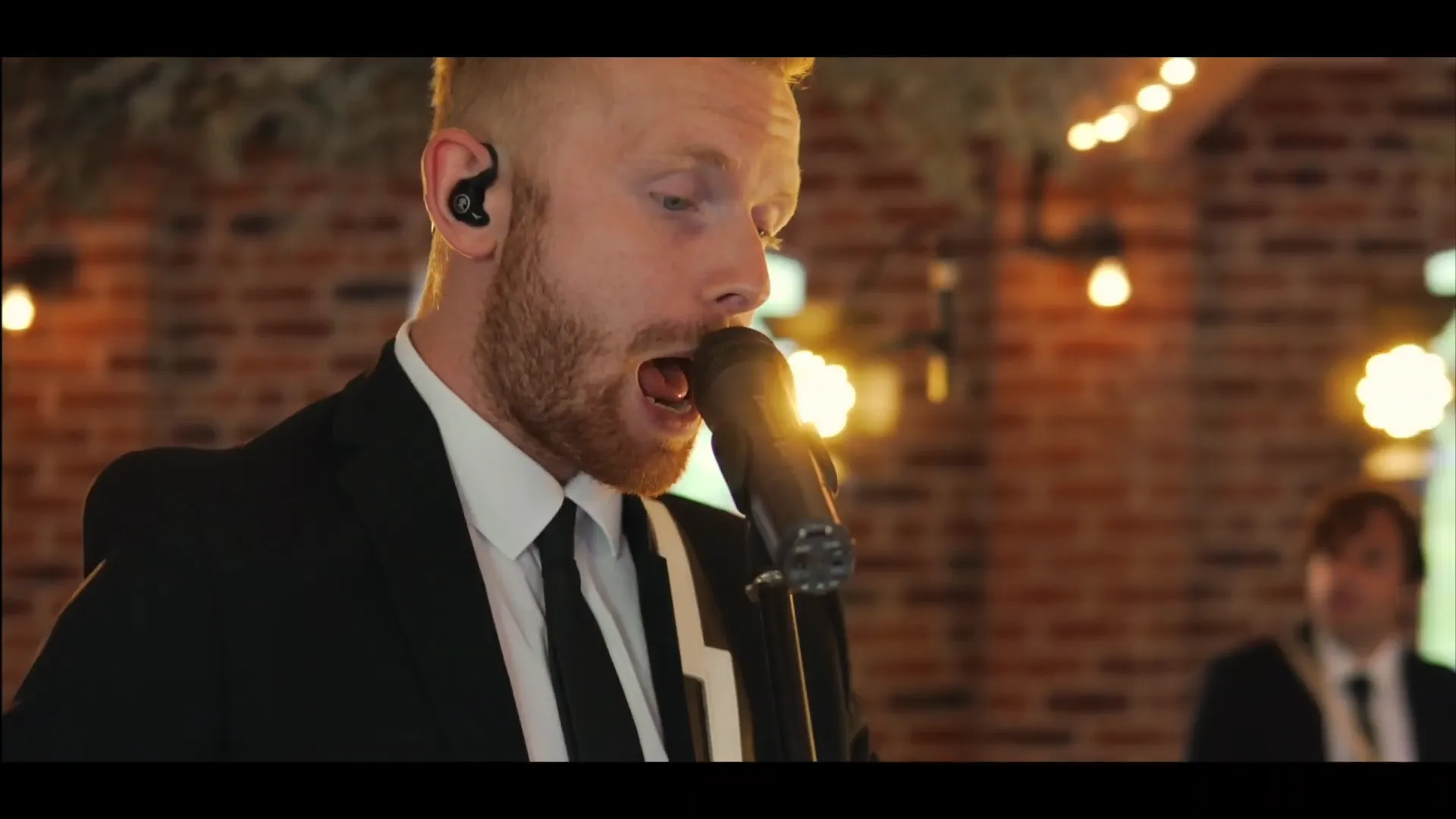 Man in suit singing into a microphone with brick wall and string lights in the background.