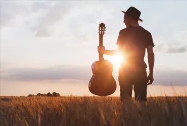 Person wearing a hat holding a guitar in a field with the sun setting behind them.