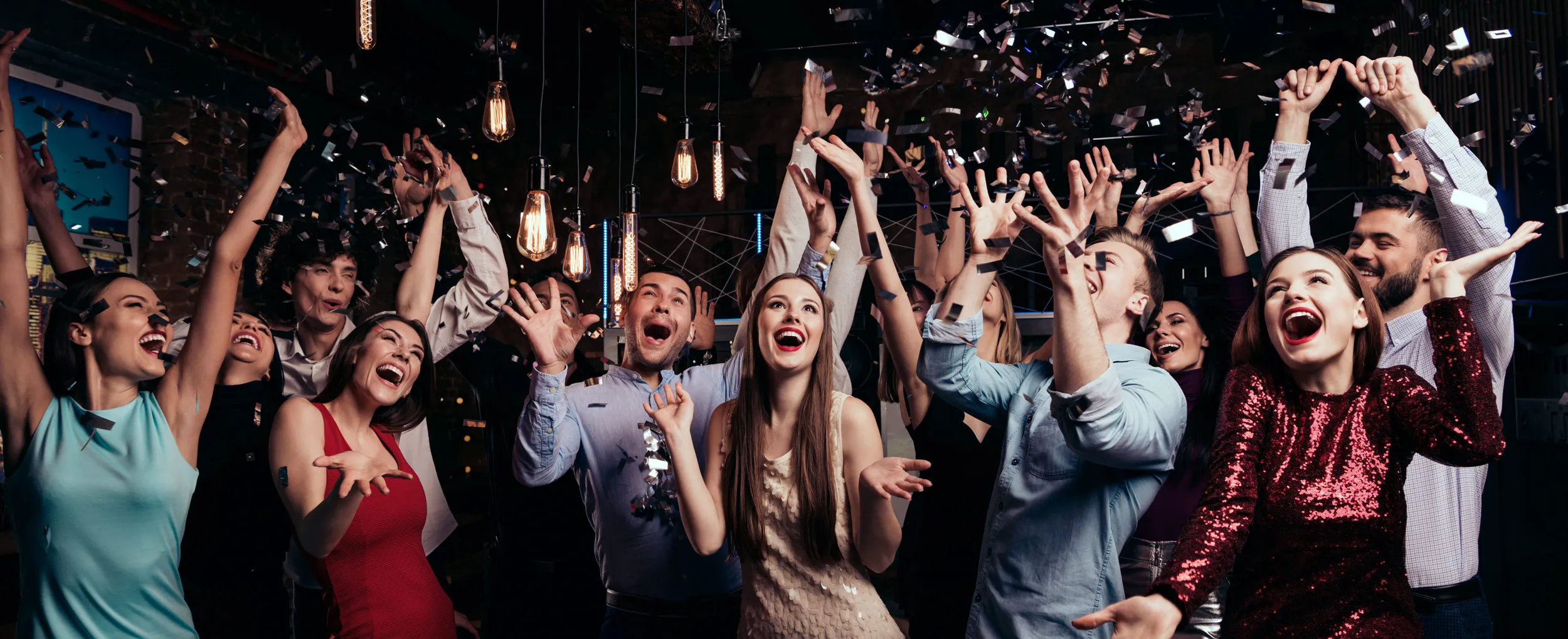 Group of young adults joyfully celebrating with raised arms and confetti falling around them at a festive party.