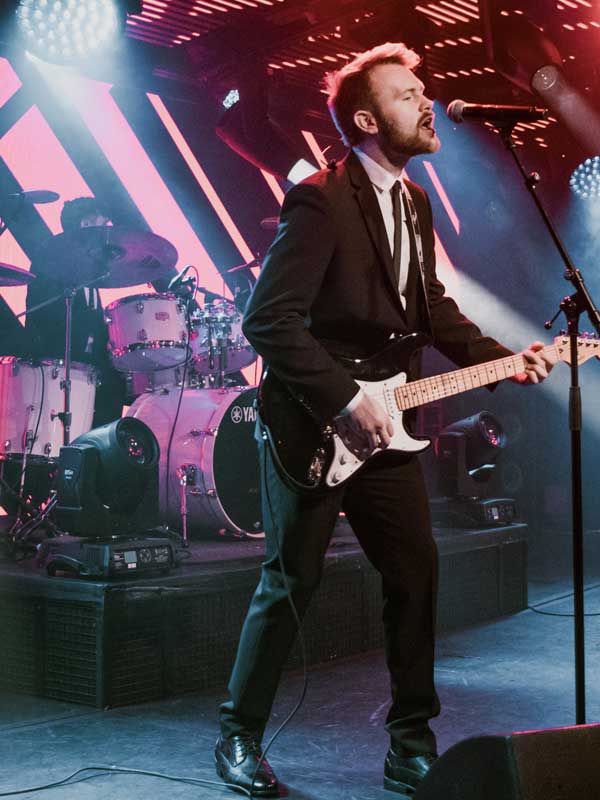 Man in suit playing electric guitar and singing on stage with drummer and drum set in the background under colorful stage lights.