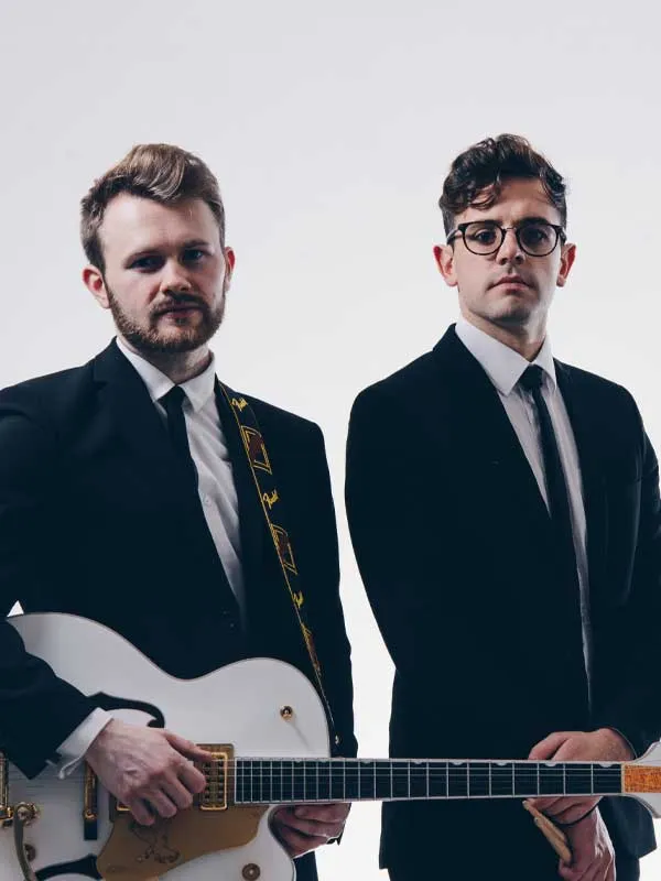 Two men in black suits and ties; one holds a white electric guitar with a patterned strap, the other stands with hands clasped.