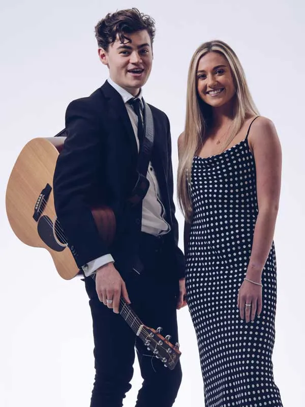 Young man in a suit holding an acoustic guitar and smiling next to a woman in a black and white polka dot dress.