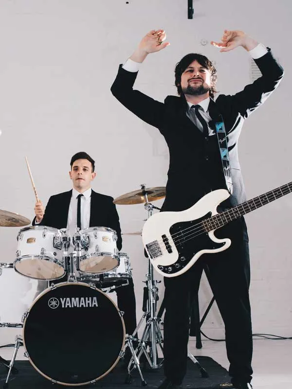Two men in black suits playing musical instruments, one on drums and the other holding a white electric bass guitar with arms raised.