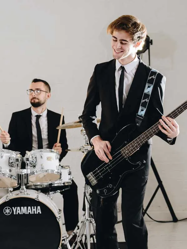 Two musicians in black suits, one playing a black bass guitar and smiling, the other playing Yamaha drums in the background.