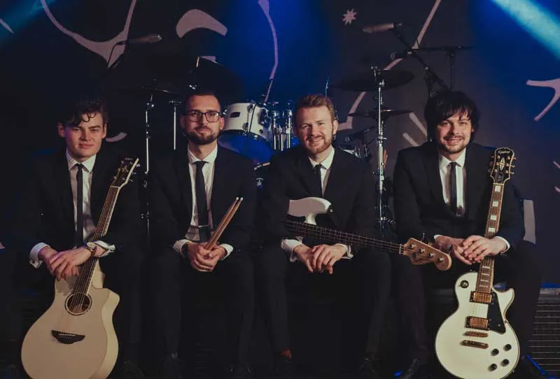 Four male musicians in suits sitting on stage with guitars and drumsticks, smiling at the camera with drum set behind them.