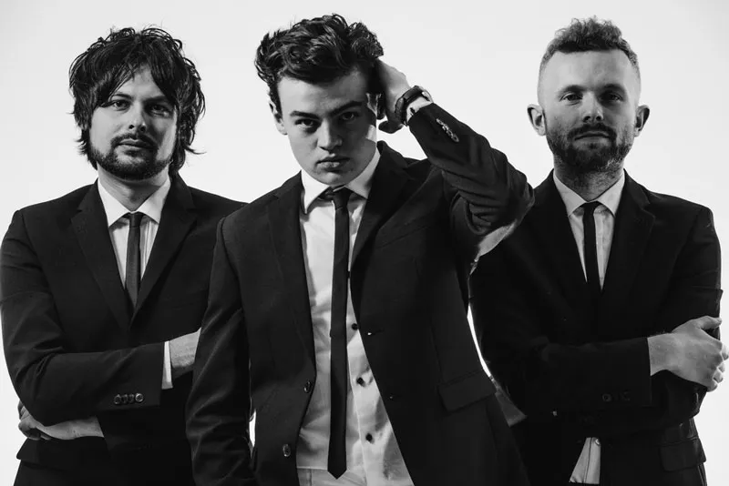 Three men in black suits and ties posing against a plain background, with the man in the center running a hand through his hair.