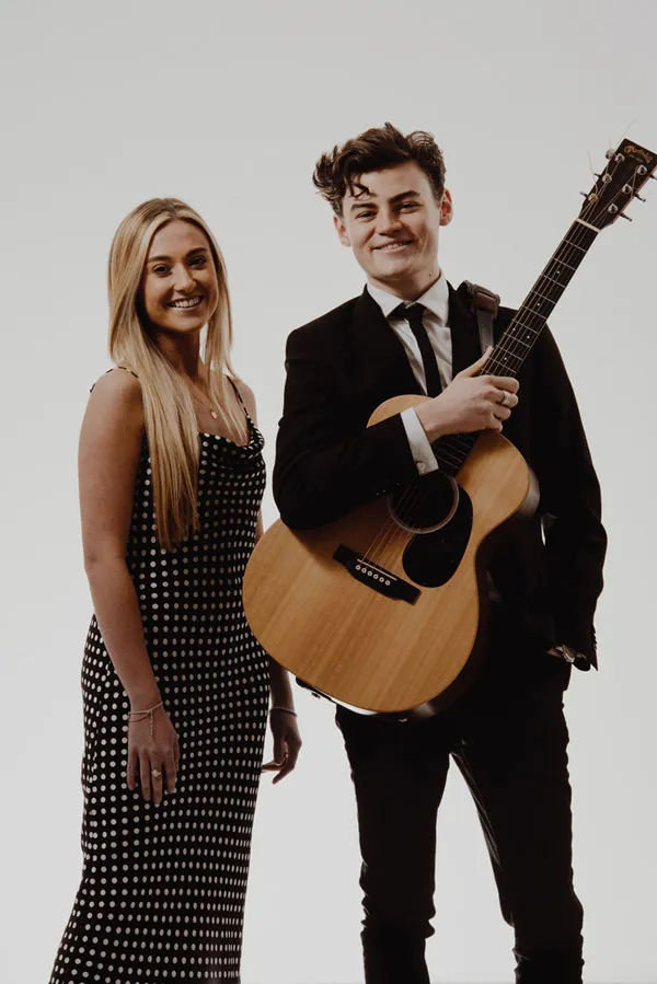 Smiling young woman in a polka dot dress standing next to a man in a black suit playing an acoustic guitar.