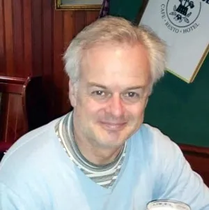 Smiling middle-aged man with gray hair wearing a light blue sweater over a striped shirt in a wood-paneled room.