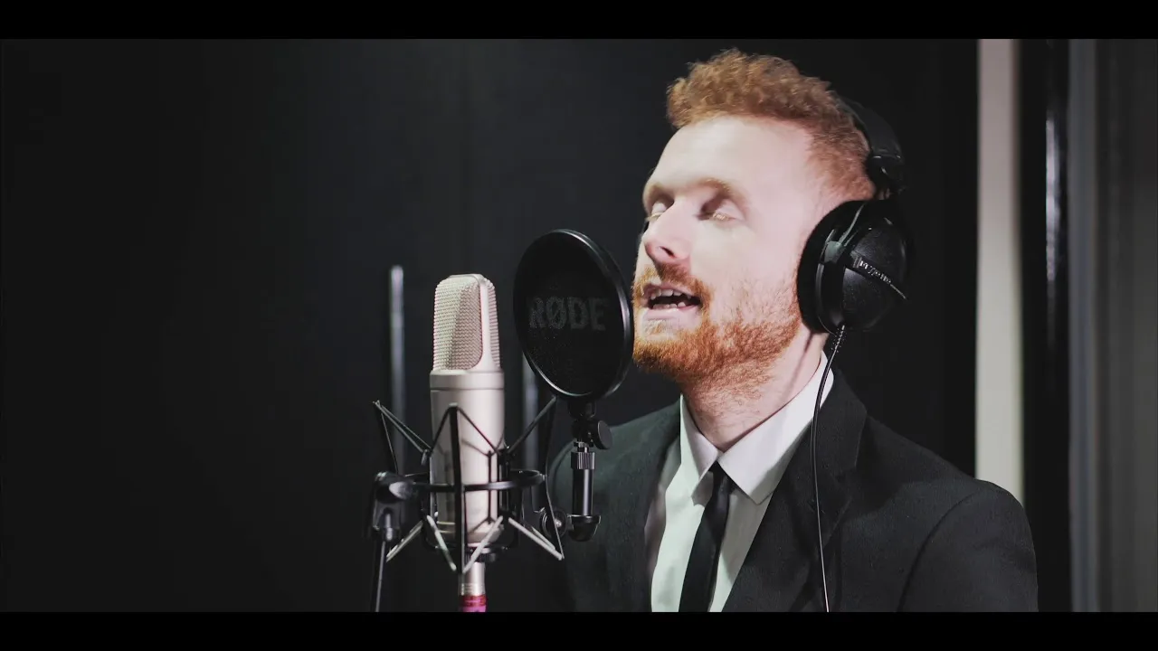 Man with red hair and beard wearing headphones and a suit singing into a microphone in a studio.