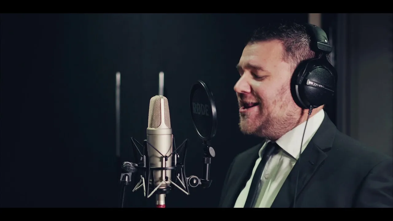 Man in suit and tie wearing headphones singing into a studio microphone with a pop filter in a recording studio.