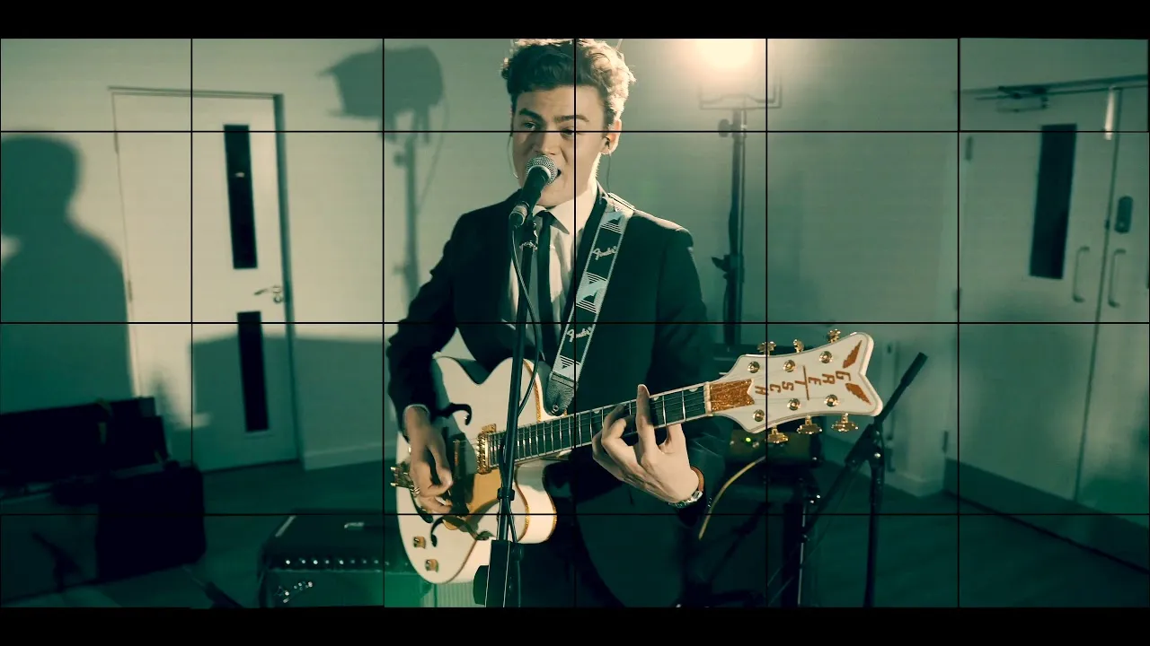 Man in a suit playing a white electric guitar and singing into a microphone in a studio setting with lighting equipment.