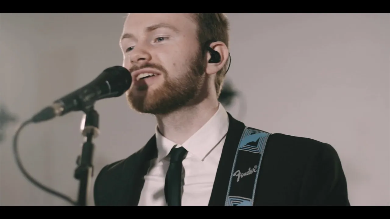 Bearded man in a suit singing into a microphone while playing a guitar with a Fender strap.