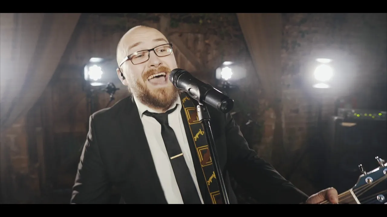 Bearded man wearing glasses and a suit singing into a microphone while playing a guitar on stage with bright lights behind him.