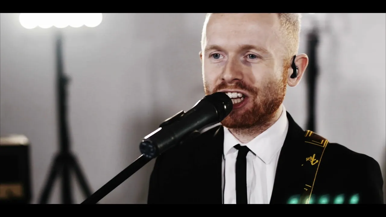 Close-up of a man with red hair and beard singing into a microphone, wearing a black suit and white shirt with a tie, and an in-ear monitor.