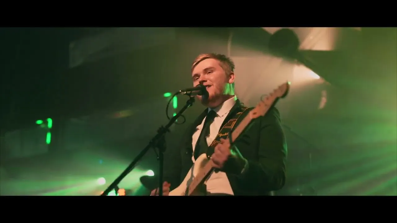 Young man in a suit playing an electric guitar and singing into a microphone on a dimly lit stage with green lights.