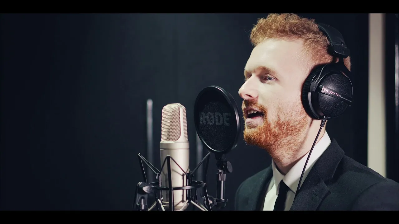 Man with red beard and curly hair wearing headphones and a suit singing into a studio microphone.