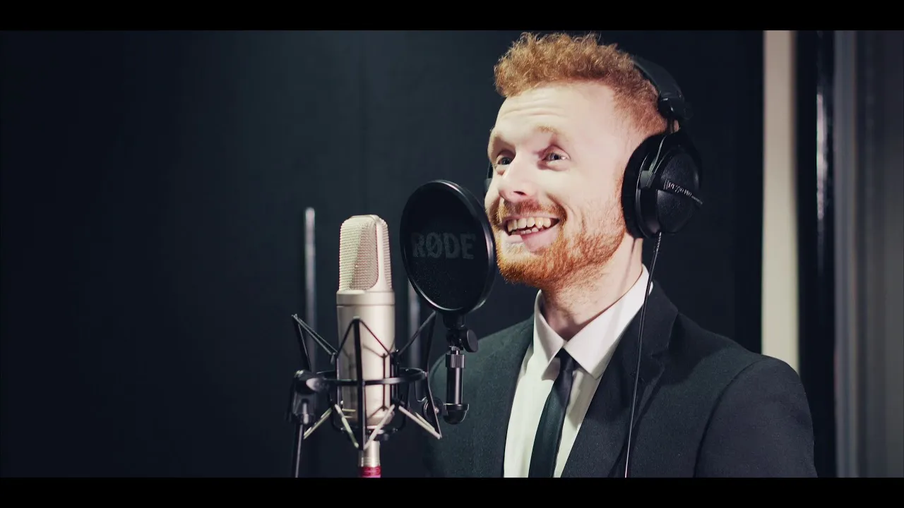 Young man with red hair and beard wearing headphones and a suit, speaking into a studio microphone.