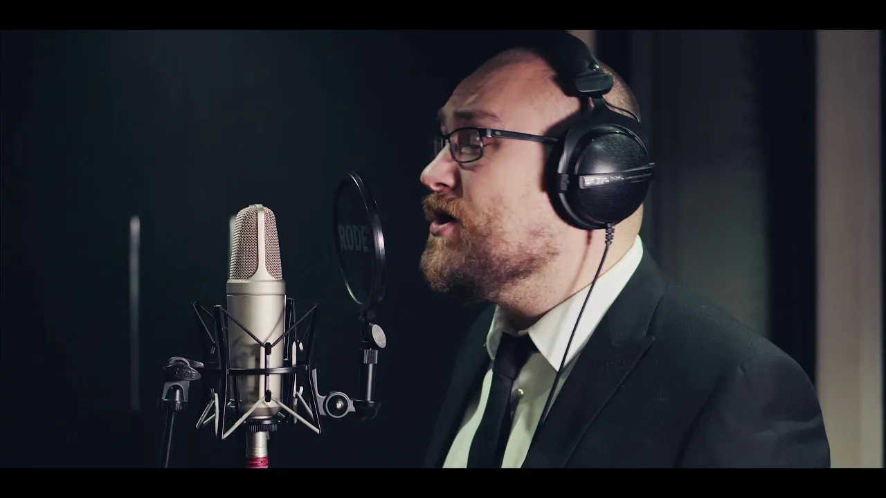 Bearded man wearing glasses and headphones singing into a studio microphone with pop filter.
