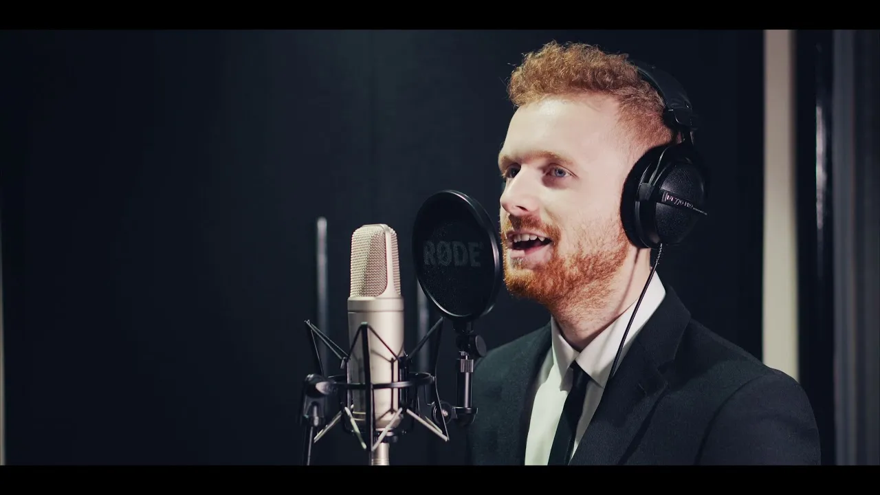 Man with red hair and beard wearing headphones speaking into a studio microphone in a recording booth.