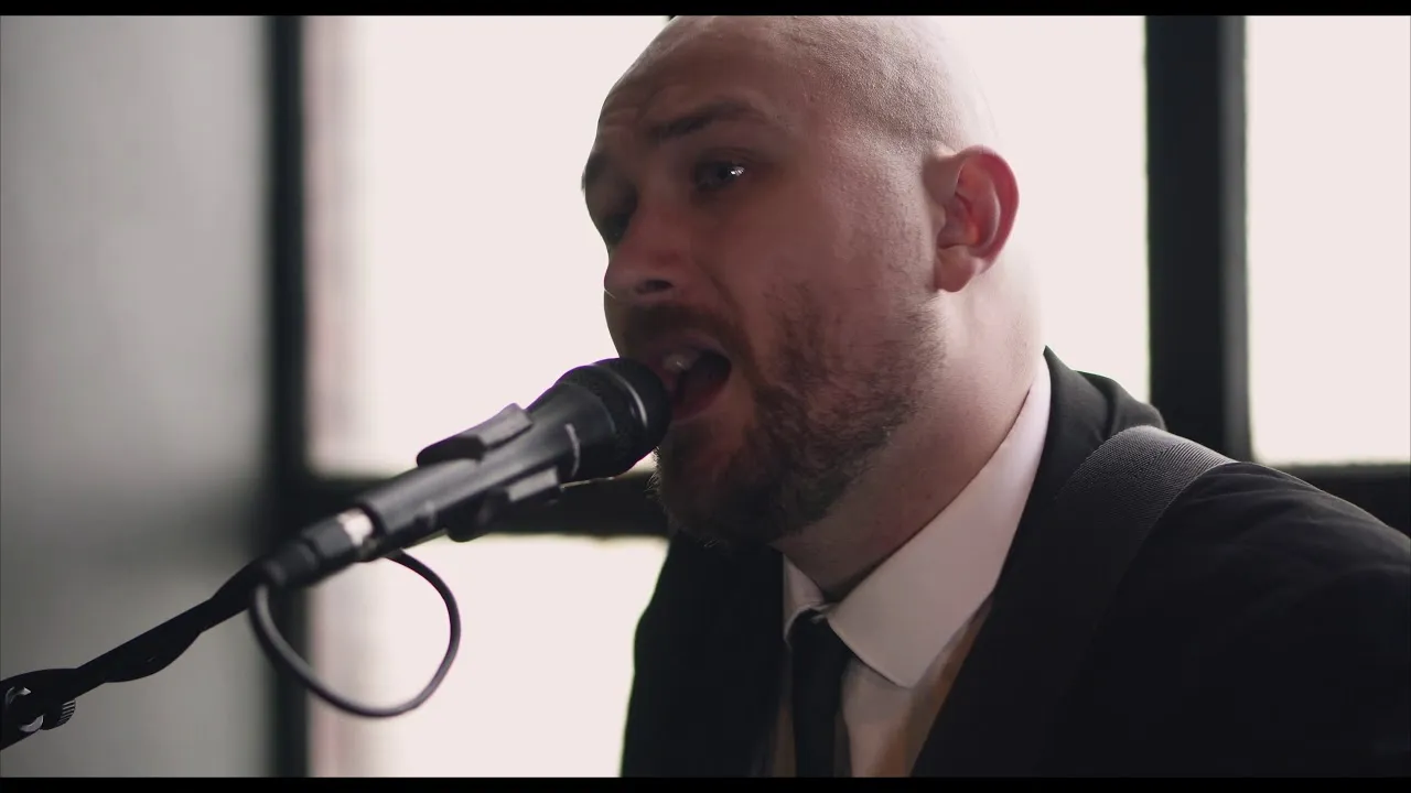 Bald man with a beard singing into a microphone indoors near a window.