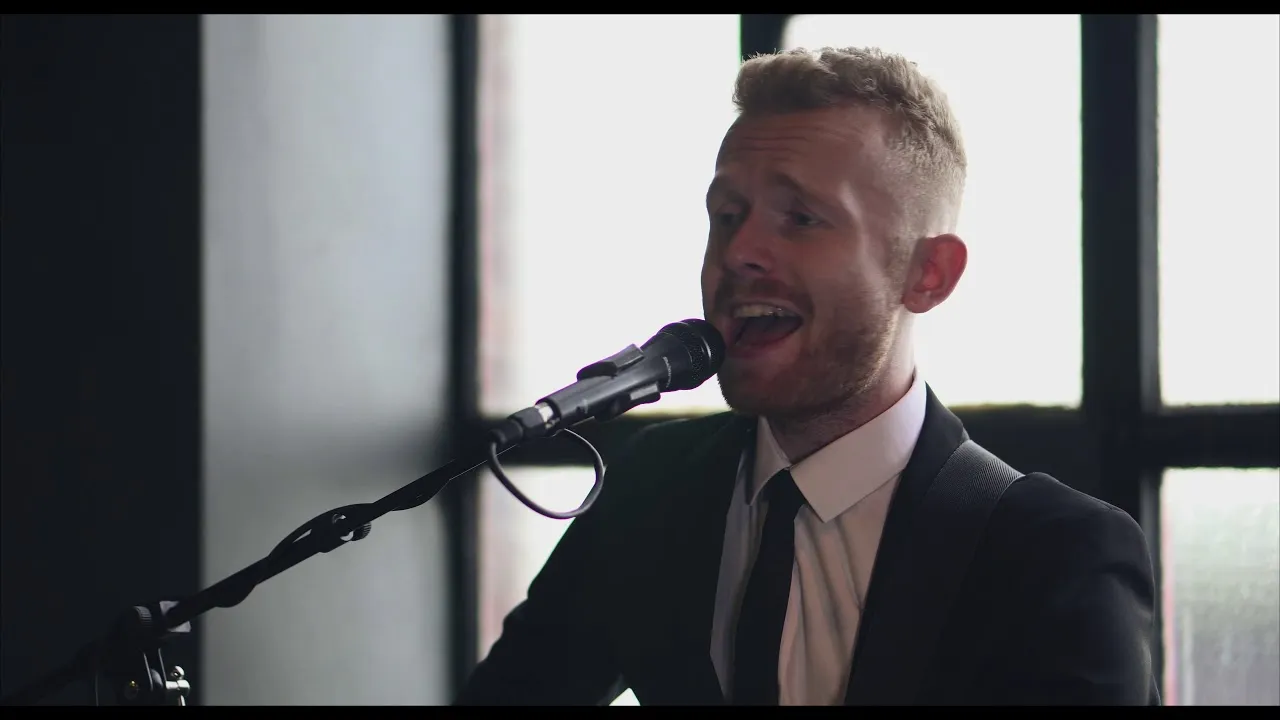 Man in a suit singing into a microphone in front of a window.
