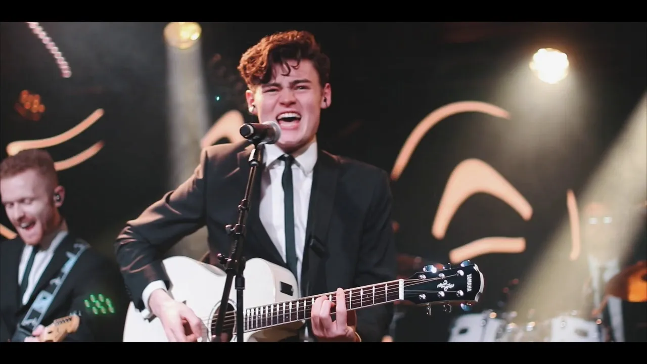 Male musician singing passionately and playing white acoustic guitar on stage with band members in the background.