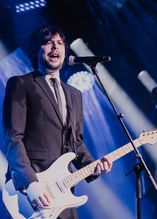 Male musician in a suit singing into a microphone while playing a white electric guitar on stage with blue spotlights.