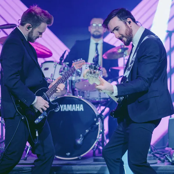 Two male guitarists in suits playing electric guitars on stage with a drummer in the background behind a Yamaha drum set.