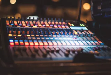 Close-up of an audio mixing console with illuminated buttons and sliders in a dim environment.