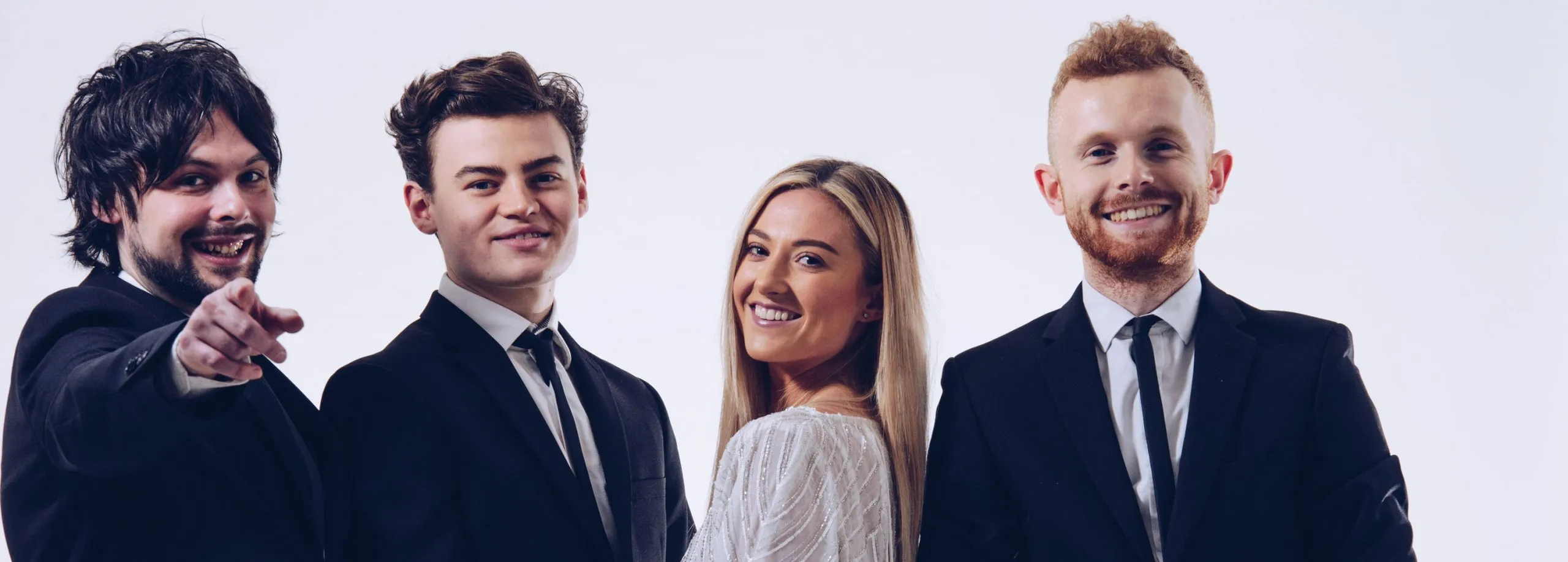 Group portrait of four smiling young adults dressed in formal attire against a white background, with one man pointing toward the camera.